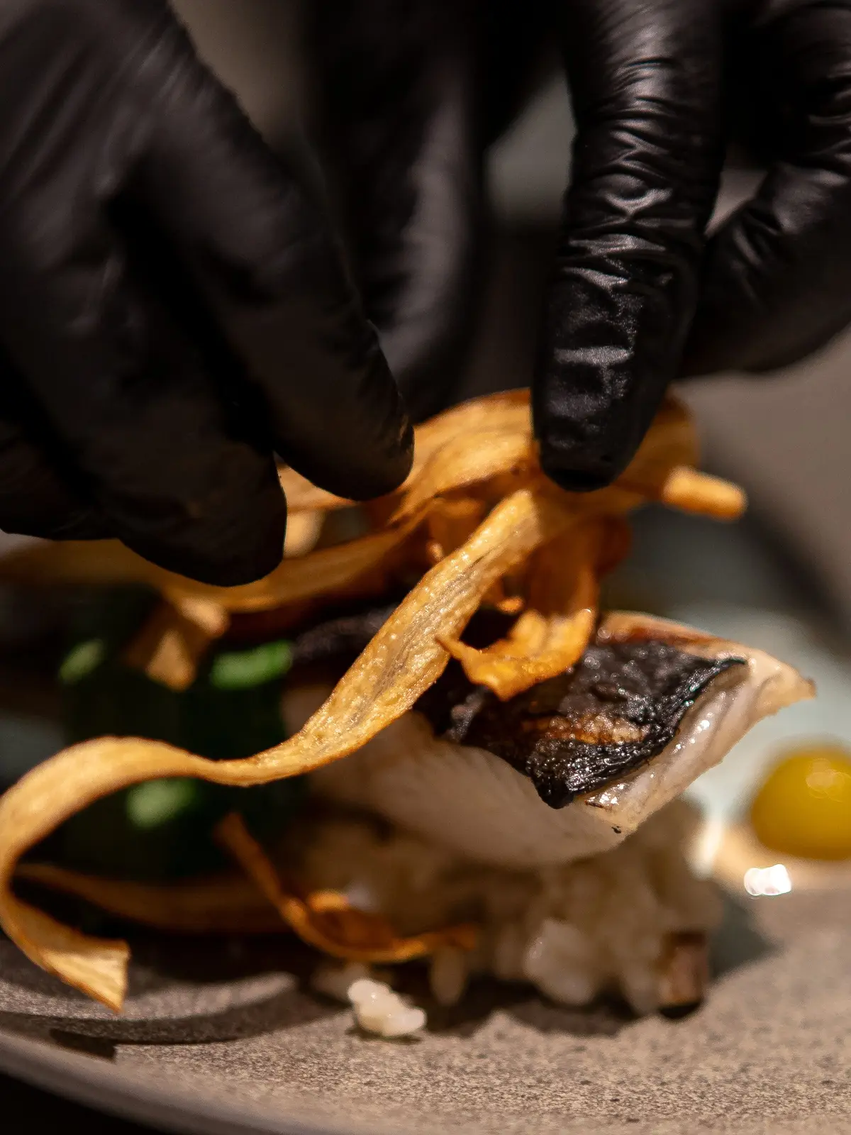 Des mains gantées de noir déposent des chips de légumes sur un poisson à la peau grillée