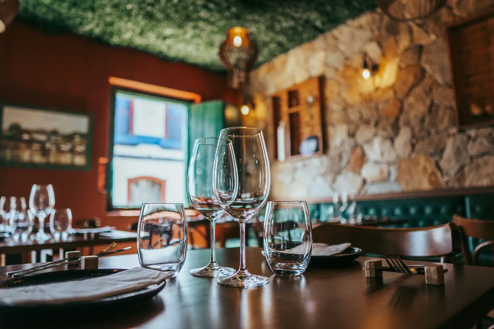 La salle de L’Ardoise de Camille : verres à pied sur une table dressée, mur de pierre éclairé par des appliques et banquette verte
