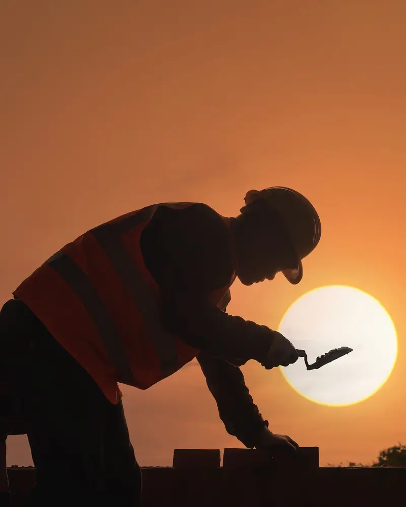 Silhouette d'un maçon casqué posant une brique à la truelle, à contre-jour au coucher du soleil (photo d'illustration pour Thomas Ravel)